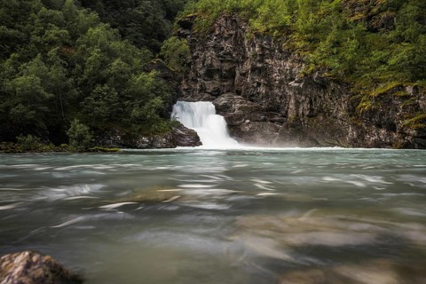 A peaceful waterfall cascades into a crystal-clear river surrounded by lush green forests and rocky