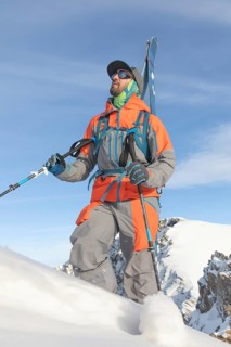 A skier in vibrant gear with skis strapped, climbs a snowy mountain. The clear blue sky and