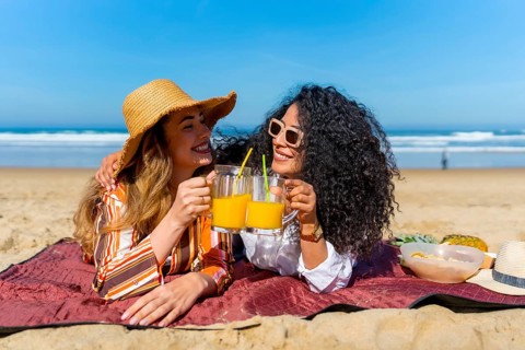 Two cheerful women friends enjoying a summer picnic on a sandy beach, raising glasses of orange