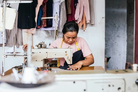 A asian seamstress is intently sewing in a machine, surrounded by hanging garments in a cozy tailor