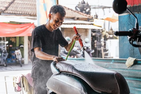 A motorcycle cleaner is smiling as he uses a pressure hose to wash a bike seat. The setting is a