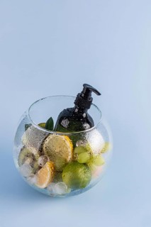 A black pump bottle of shower gel is surrounded by fresh citrus fruits and ice in a glass bowl