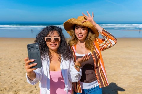 Two women friends smiling and posing for a selfie using a smartphone on a sandy beach with the