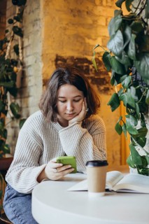 A young woman in a cozy sweater sits at a bookstore cafe table, scrolling on her smartphone An open