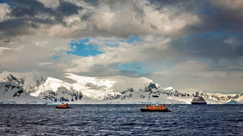 Two inflatable boats and a cruise ship in front of snowy mountains under a cloud cover at sea,