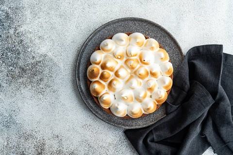 A lemon cake topped with toasted meringue sits on a dark plate, complemented by a cloth. The pie,
