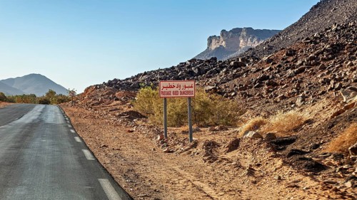 A tarmac country road runs through a dry desert landscape with a sign and mountains in the