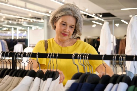 Mature woman browsing through clothing on a rack in a modern store. Her gray hair and elegant