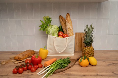 A reusable tote filled with fresh produce and baguettes sits on a wooden table. Surrounded by