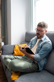A man in casual attire sits cross-legged on a gray sofa, writing in a notebook. The living room is