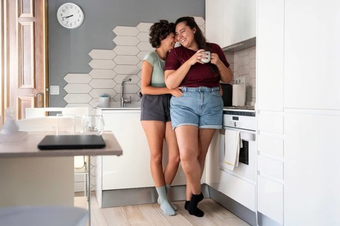 A happy lesbian couple shares a playful moment in a modern kitchen. One partner sits on the