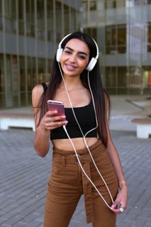 Young woman listening to music on headphones, holding her smartphone. She is outdoors, wearing