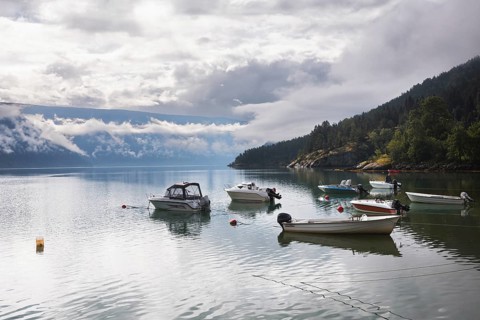 A serene lake in Norway reflects moored boats, surrounded by misty mountains and lush greenery The