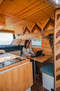 A woman works remotely on her laptop inside a cozy camper van, parked with a scenic view outside.