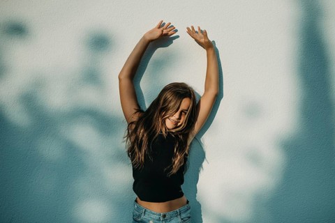 A teen girl stands with arms raised against a wall, with a joyful expression and shadows from tree