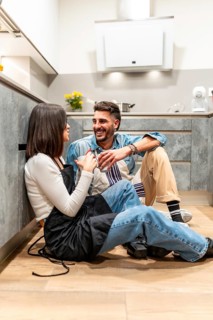 Two chefs are sitting on the floor of a modern kitchen, wearing aprons and enjoying a coffee break