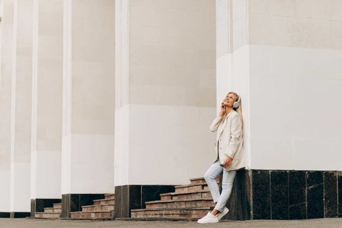 A businesswoman leans against a building column, enjoying music through headphones. Dressed