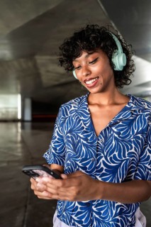Woman with curly hair wearing blue patterned shirt listens to music on headphones while smiling and