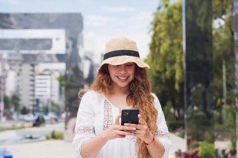A young American Latin woman smiles while using her smartphone outdoors. Dressed in casual attire