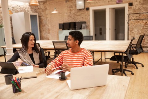 Two professionals collaborate in a modern coworking space, discussing ideas with laptops and