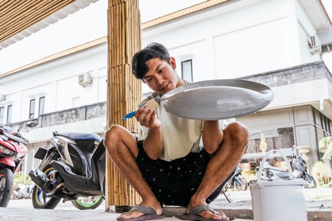 A Balinese artist skillfully paints a sculpture outdoors, surrounded by local architecture and