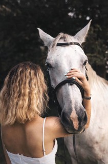 A young woman with wavy blonde hair embraces a white horse in a serene outdoor setting. The image