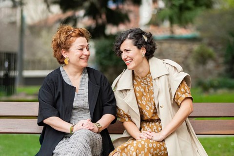 Two women sit on a park bench, engaged in friendly conversation and laughter. The vibrant colors of