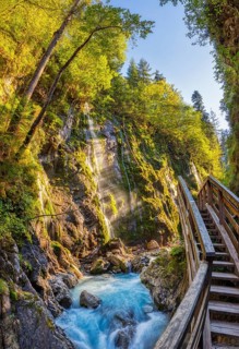 An idyllic hiking trail with a wooden staircase leads along a waterfall in the middle of a green