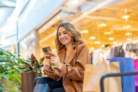 Young woman smiling and scrolling on her smartphone while resting with shopping bags in a bright