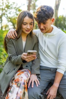 Smiling young couple sitting in a park, sharing a moment while looking at a smartphone, surrounded