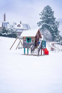 A snow-covered playhouse with swings on a snowy playground in front of wintry trees, Enzklösterle,