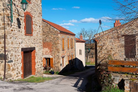 Eine ruhige Dorfstraße mit steinernen Häusern und Fensterläden unter einem sonnigen, blauen Himmel,