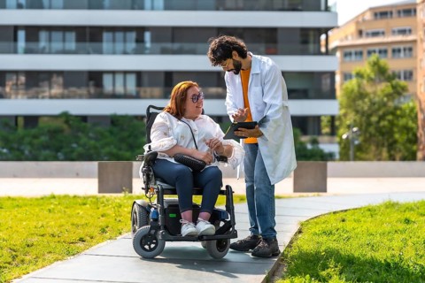 Doctor holding tablet explaining diagnosis to smiling woman sitting in wheelchair in urban park