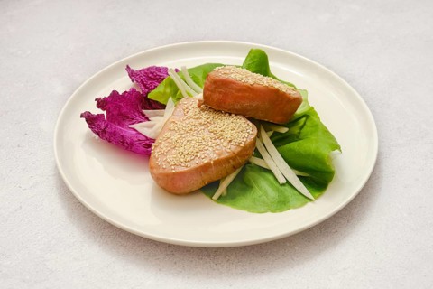 Two pieces of sesame-coated tuna fish are on a plate with a mix of vibrant greens and thinly sliced