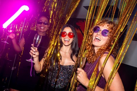 Three cheerful young women with colorful sunglasses are holding glasses of sparkling wine, enjoying