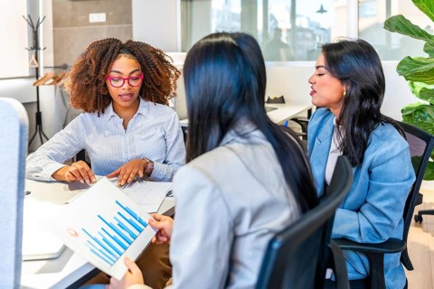Three businesswomen are discussing over financial charts sitting at a desk in a modern coworking