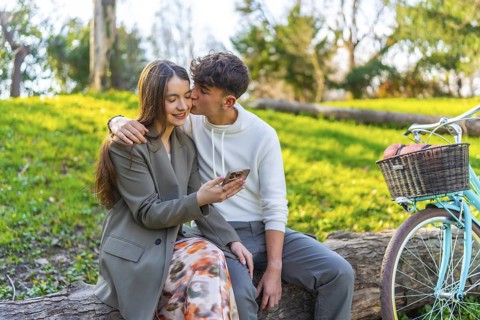 Happy couple sitting on a log in a park, sharing a moment with a smartphone, bicycle nearby,