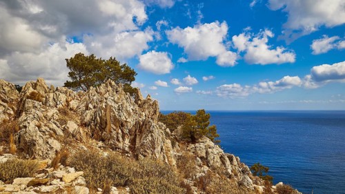 Steep rock formations with trees in front of the blue sea and a cloudy sky, near Kato Lakkos Beach,