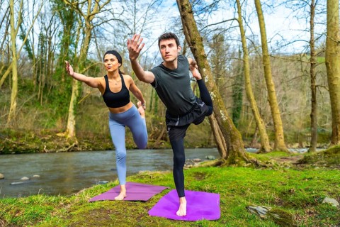 Man and woman practicing yoga in nature, performing the natarajasana pose on mats near a river,