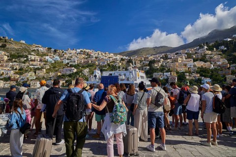 People waiting in a coastal town with colourful houses on a hill under a blue sky with clouds, Symi
