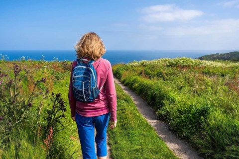 A hiker with a rucksack on a coastal path in Cornwall. View of the sea. Wildflowers in bloom. Near