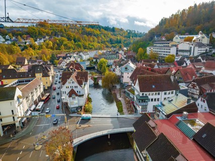 Small town with river and bridge, surrounded by autumnal trees and hills, extension of the Calw