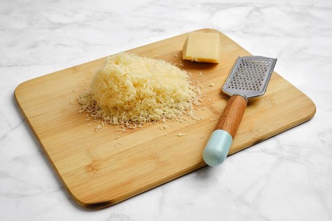 A wooden cutting board holds a pile of finely shredded cheese beside a block of cheese and a metal