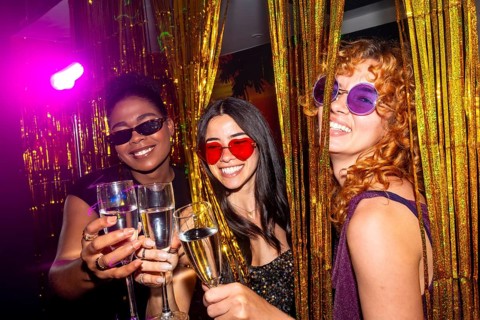 Three multi ethnic female friends are toasting with champagne flutes at a nightclub, enjoying the