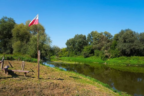 Riverbank with Polish flag and benches, surrounded by trees under a clear sky, bow River, border