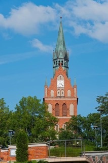 High red-brick church tower with clock, surrounded by green trees under a bright blue sky, Sacred