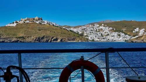 View from a ferry to a white village on a hill island under a clear sky, Chora, Astypalea,