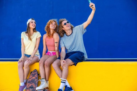 Three cheerful friends taking a selfie with a smartphone while sitting on a yellow wall with a blue