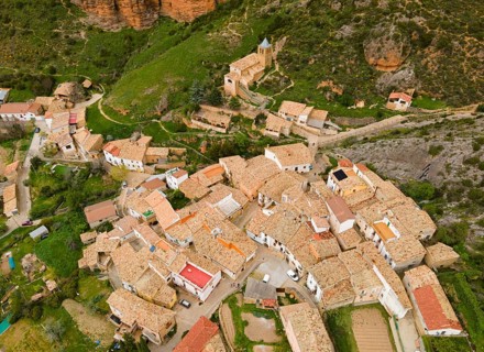 Aerial view of a village with traditional buildings nestled against a green hill with steep cliffs,