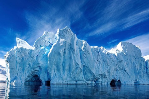 Large ice wall with caves in the Arctic is reflected in the calm water under a clear sky, icebergs
